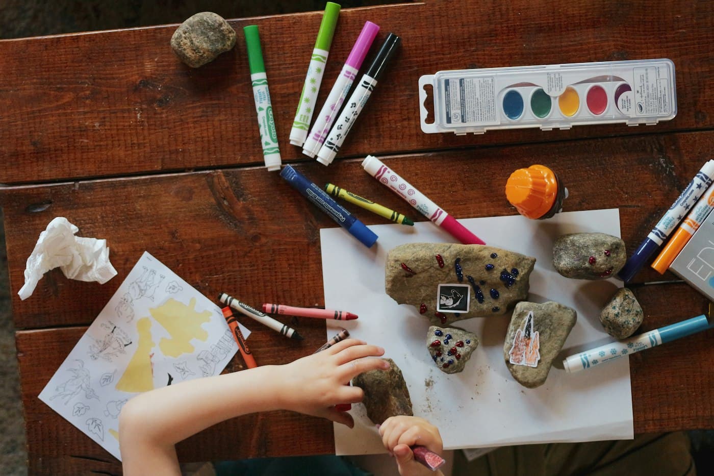 A child’s hands arranging stones, crayons and paper on a wooden table.