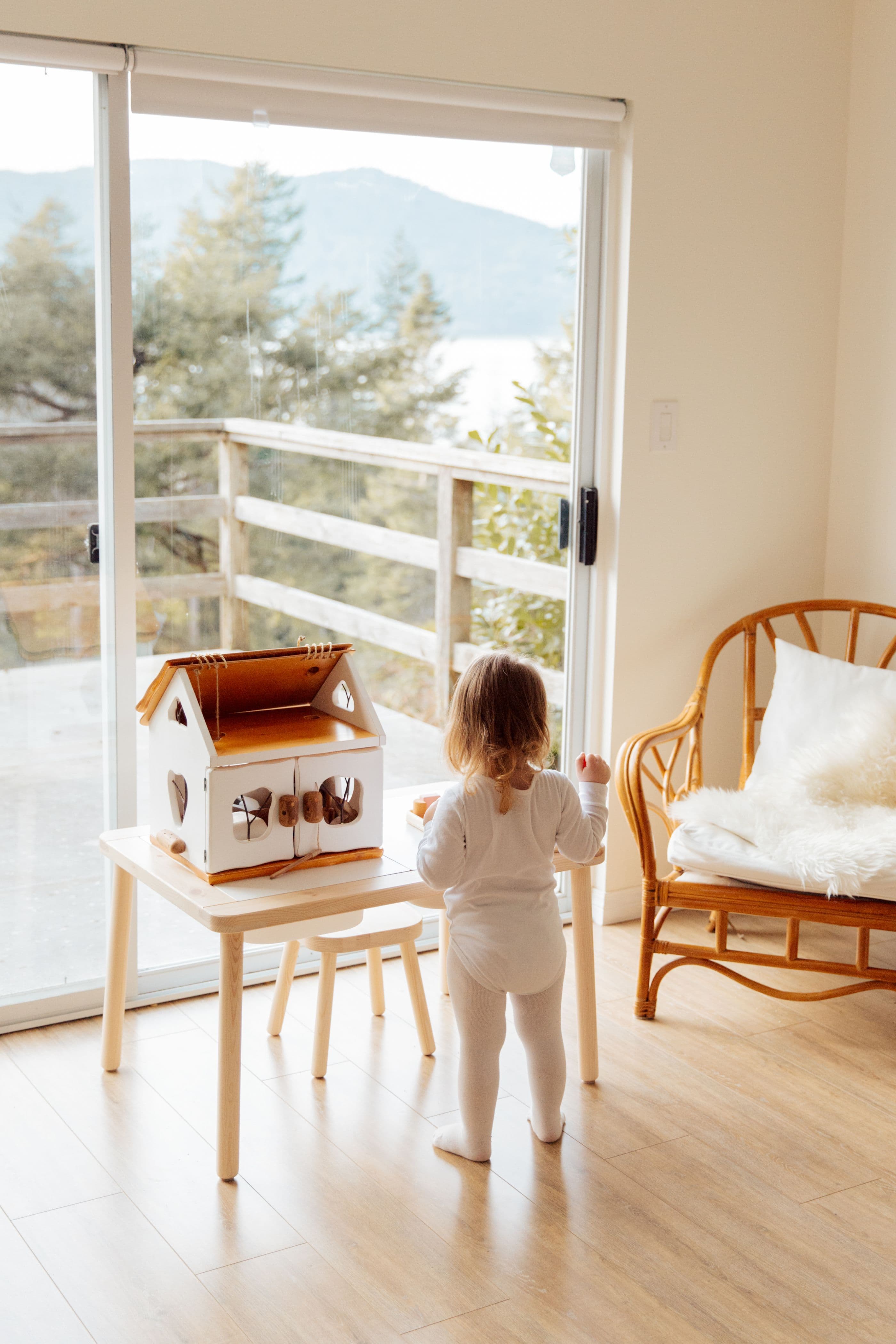 A toddler at a low table by a tall window, playing with a wooden house.