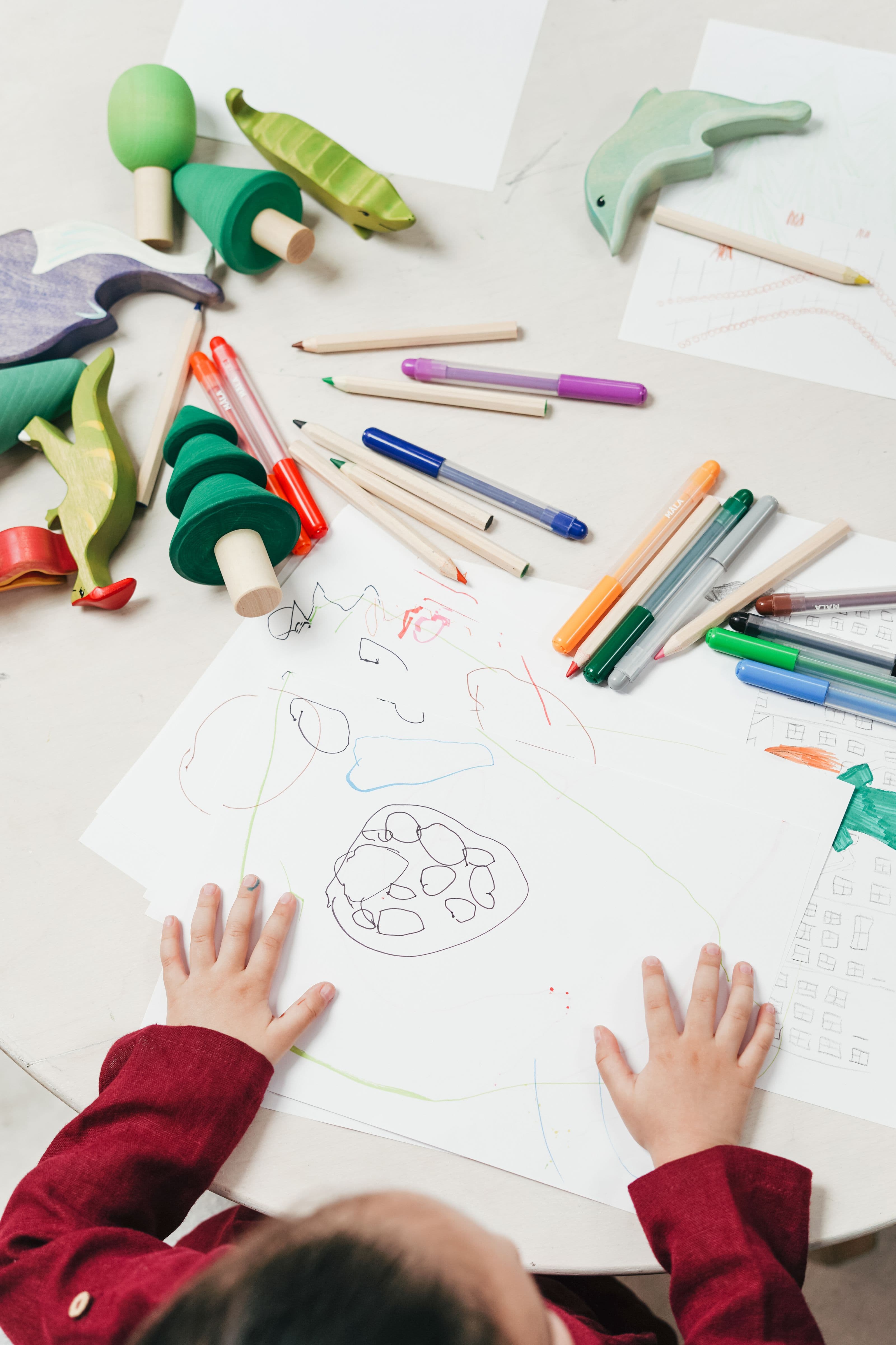 A child’s hands on a drawing, wooden toys and pencils on the table.