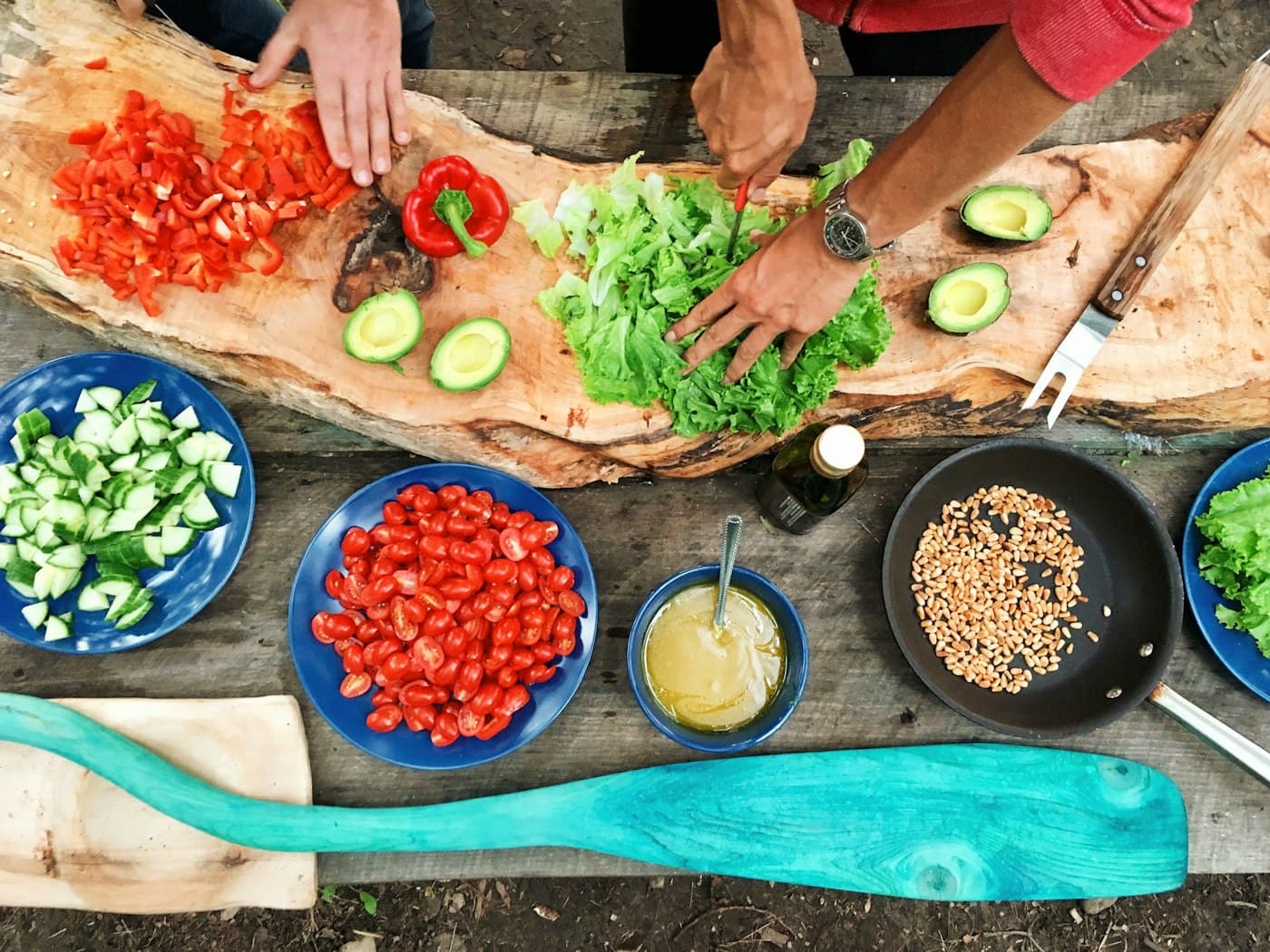 Hands preparing vegetables together on a long wooden table.